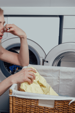 Woman reacting to bad smell from front-loading washing machine while holding laundry basket in laundry room.