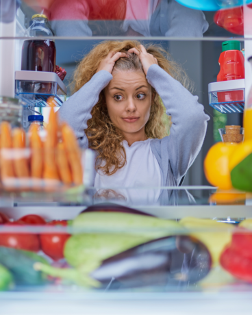 Woman looking stressed inside an overcrowded refrigerator with food items, representing simple fridge organization ideas and kitchen clutter.